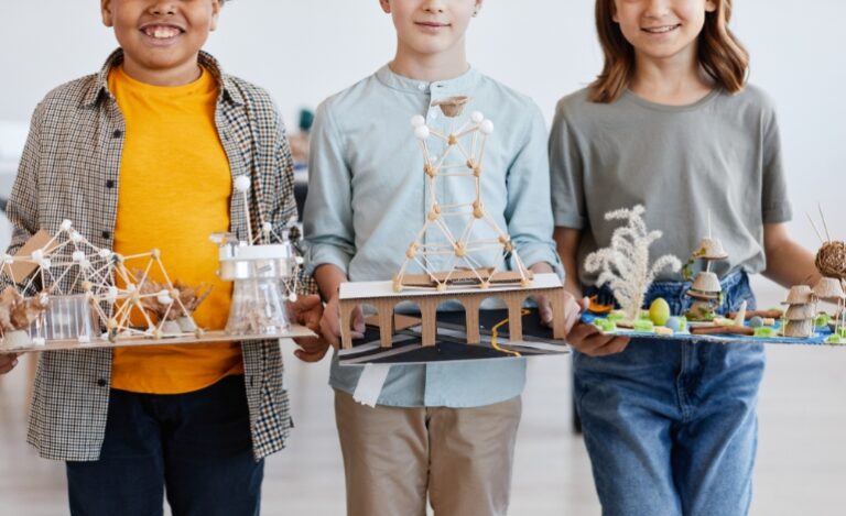 Fotografia mostrando um projeto escolar, no qual um menino com camisa xadrez azul está manuseando uma estrutura molecular feita de palitos e balas de goma.