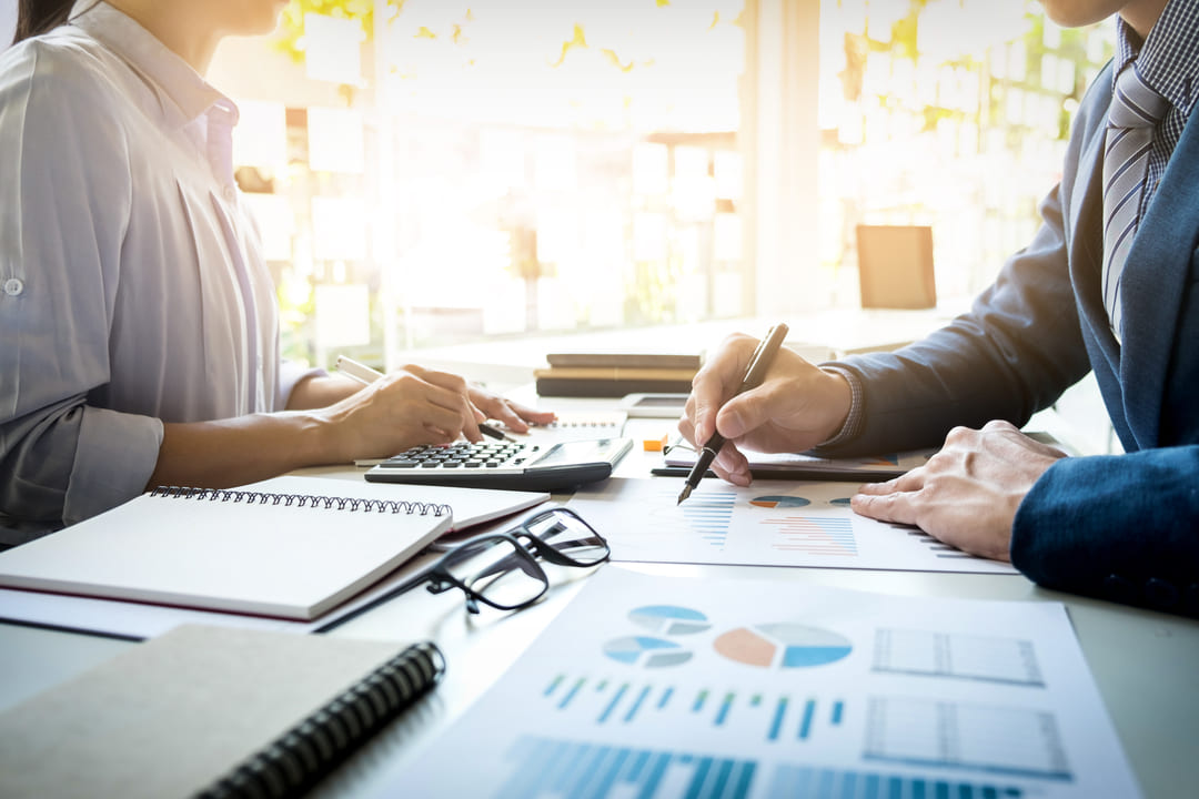 Um homem e uma mulher estão sentados em uma mesa, de frente um para o outro, enquanto analisam dados financeiros para elaborar um controle de caixa escolar