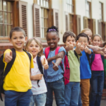 Sete crianças felizes em sua escola, posando para foto sorridentes fazendo um sinal de positivo com as mãos